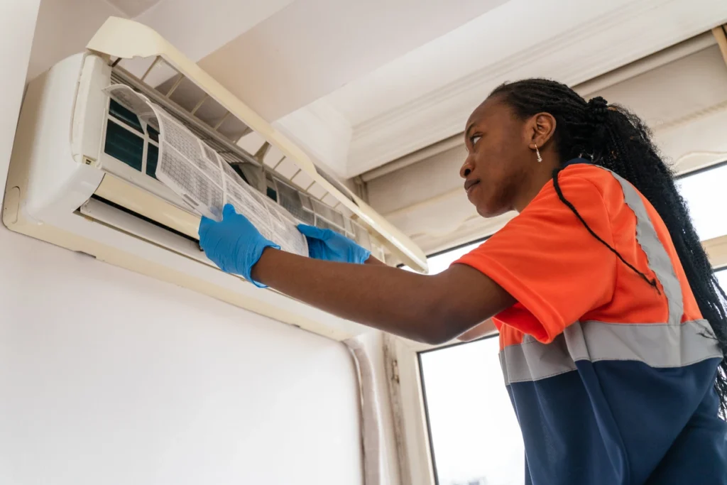 A woman in an orange shirt and blue gloves is performing maintenance on an air conditioning unit.